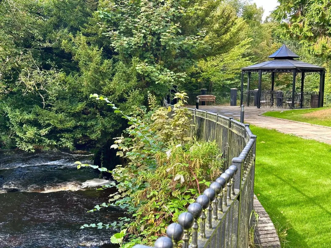 A railed path along the river through the Crossbasket estate, passing a lawn and a gazebo.