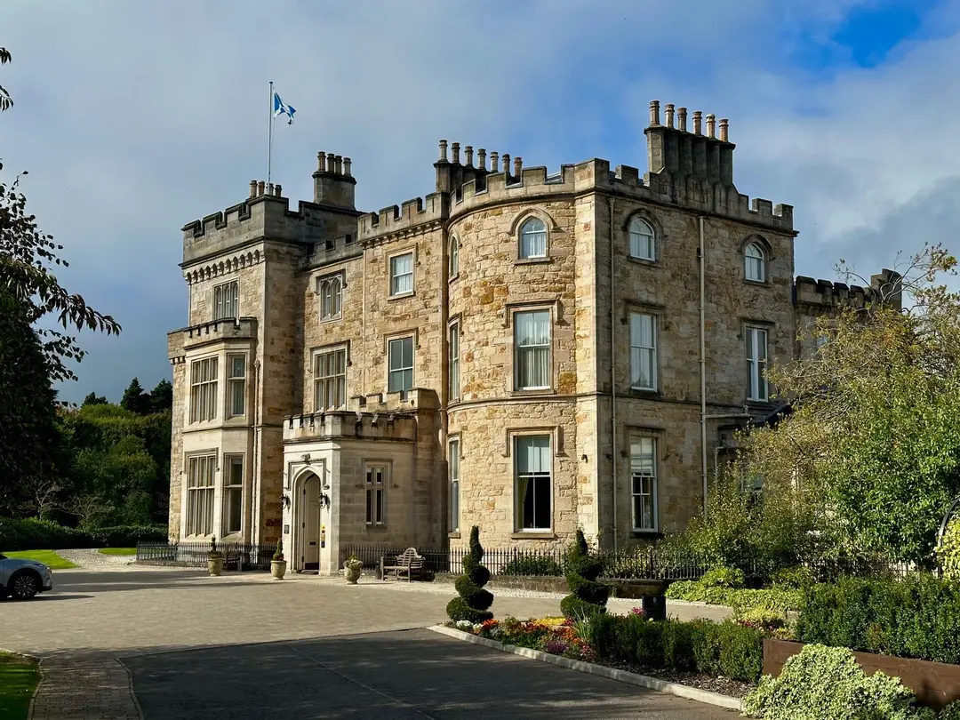 Crossbasket Castle in sunshine: a sandstone mansion with crenellated battlements and tall chimneys, a Saltire flying above