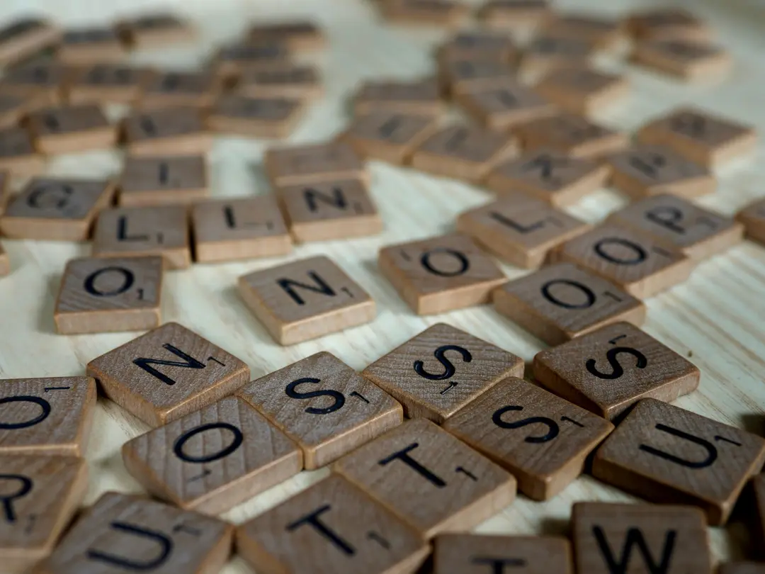 Wooden letter tiles scattered across a pale wooden table, with O, N, S, T and U in focus at the front and the rest fading out of focus behind